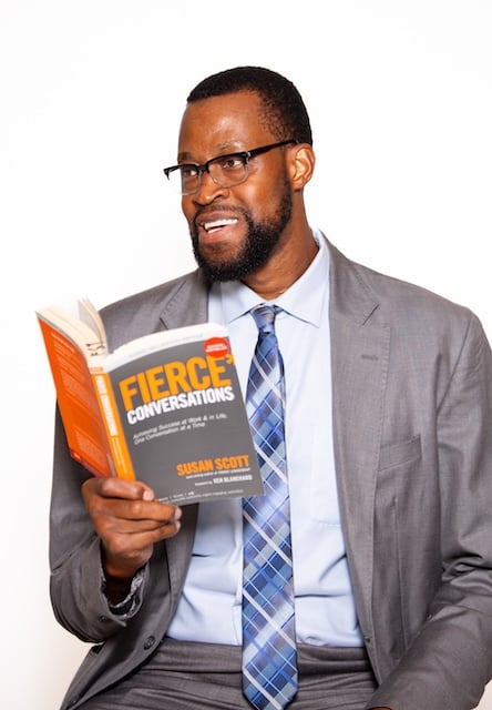 A man in a suit reads "Fierce Conversations" by Susan Scott with a smile.