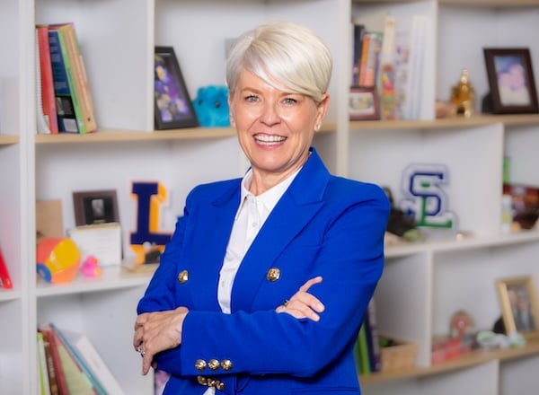Smiling woman with short white hair in a blue blazer, standing confidently with arms crossed in front of a bookshelf filled with decorative items.