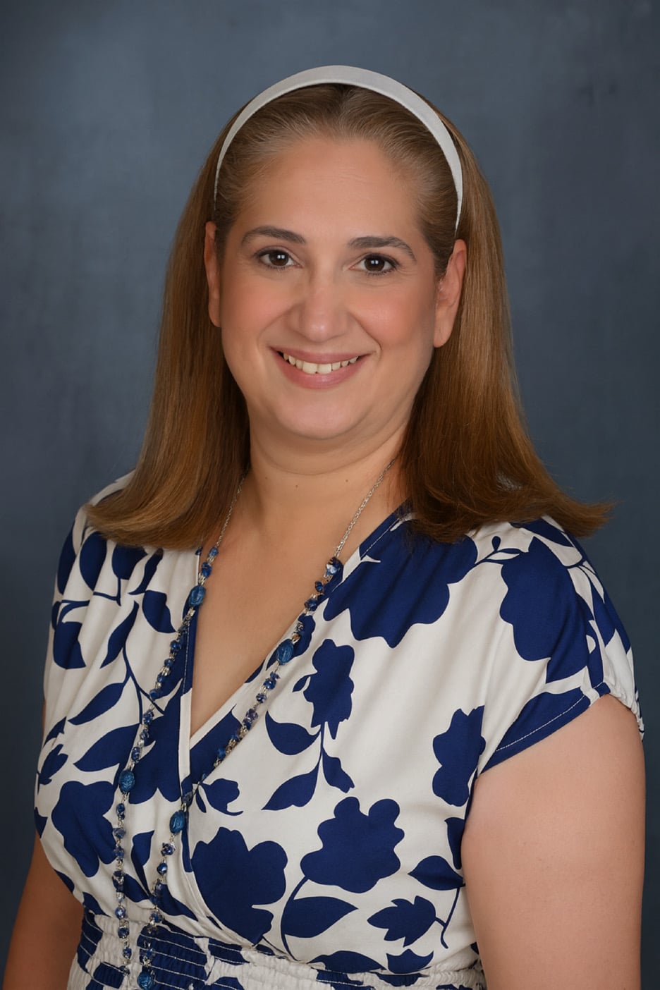 A smiling woman with medium-length brown hair wearing a floral-patterned blouse and a headband.