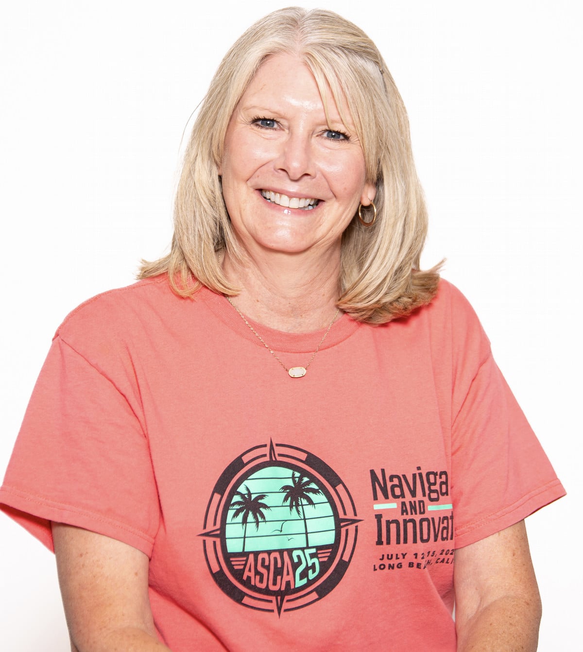 A smiling woman wearing a coral t-shirt featuring a logo for ASCA 25, which includes palm trees and the text "Navigating and Innovating."