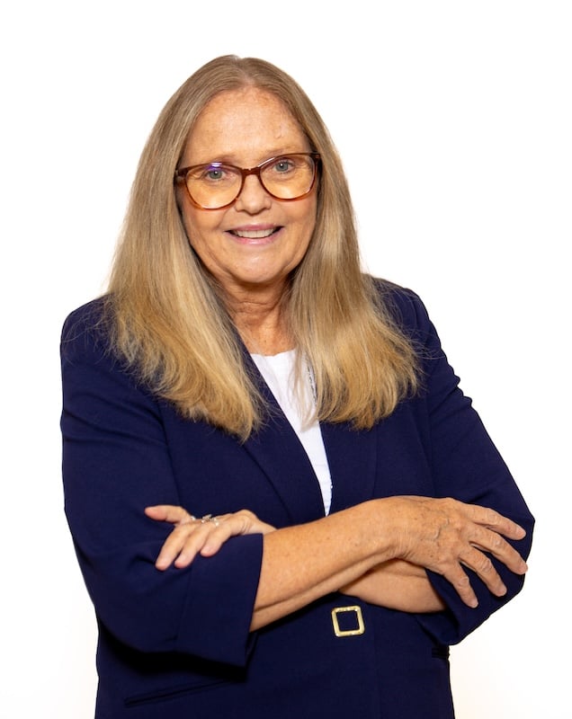 Studio portrait of a smiling older woman with long gray-blonde hair and glasses, wearing a navy blazer with arms crossed against a white background.
