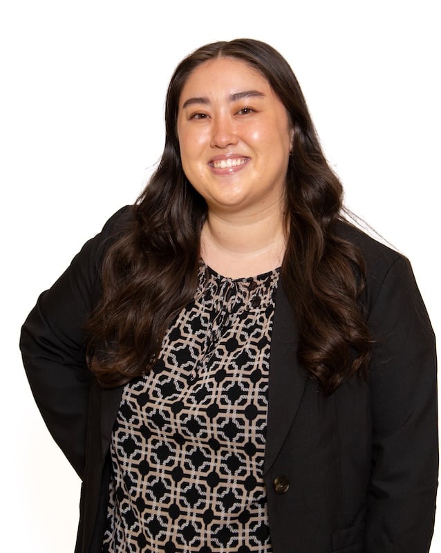 Smiling woman with long dark hair wearing a black blazer over a patterned blouse, posed against a white background.
